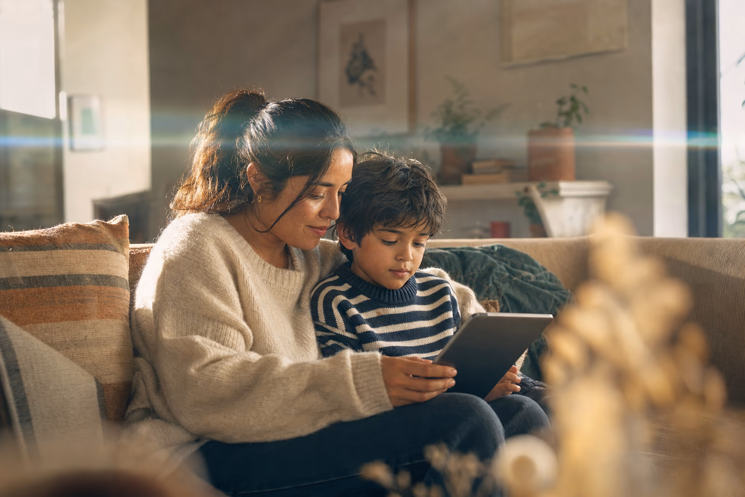 A mother and her young son sit together on a sofa at home, looking at a tablet.